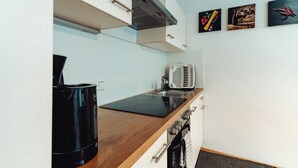 Une cuisine avec une plaque de cuisson à induction noire, un four blanc et une machine à café noire. Trois tableaux sont accrochés au mur : ils représentent des fruits, un oiseau et des piments.