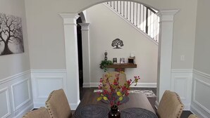Een eetkamer met een houten tafel, beigekleurige stoelen met bekleding, een vaas met bloemen en een trap op de achtergrond.