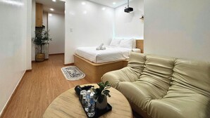 Una habitación de hotel moderna con una cama, un sofá de cuero, una mesita con una planta y botellas, y un pasillo que conduce a otra habitación.
