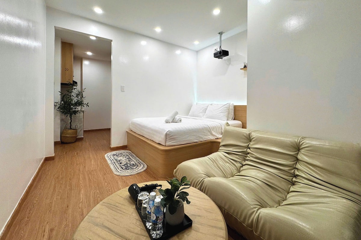 A modern hotel room with a bed, a leather sofa, a small table with a plant and bottles, and a hallway leading to another room.