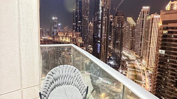 Un balcón con vista a un paisaje urbano nocturno, con edificios altos y barandilla de vidrio.
