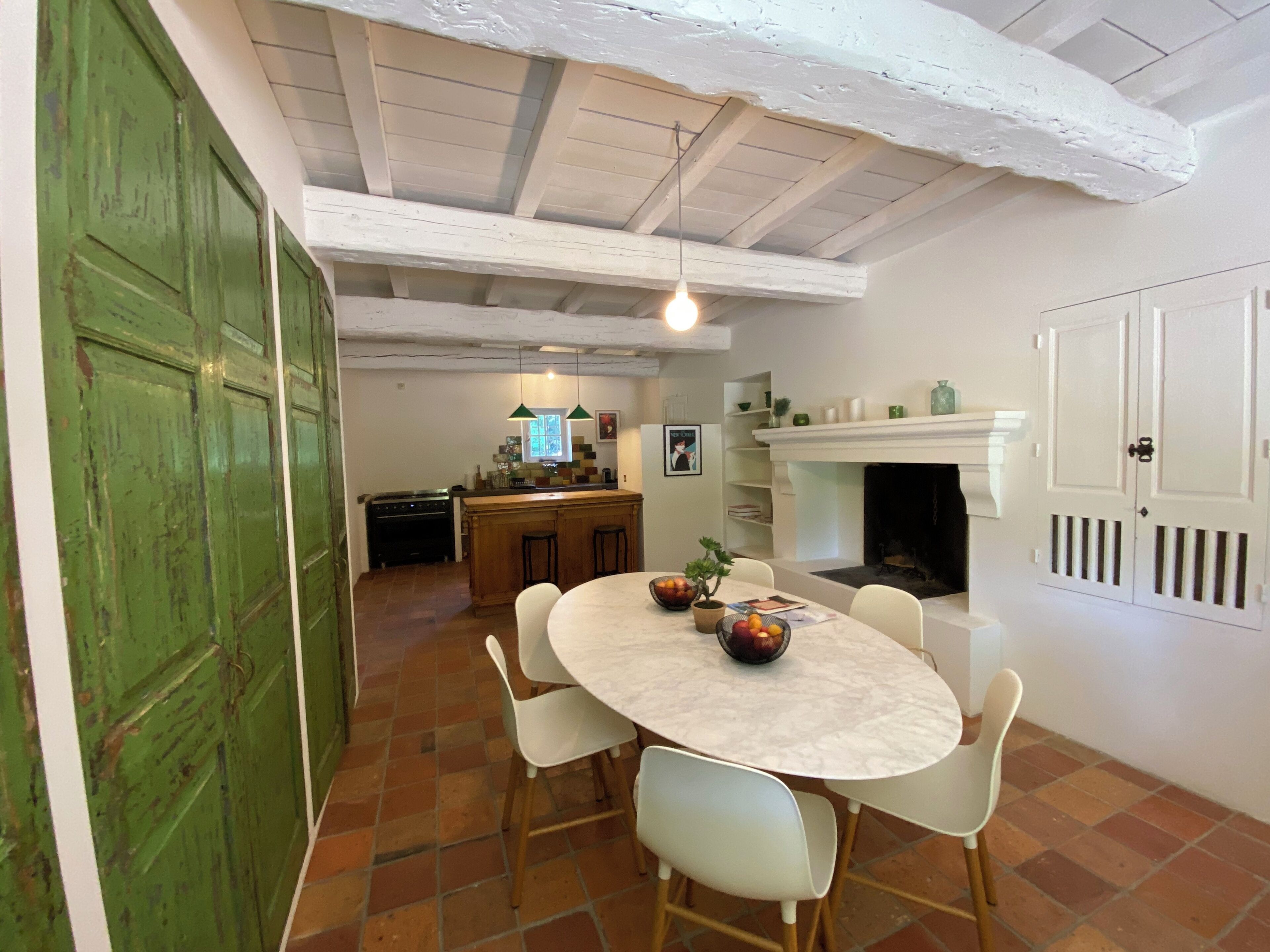 A dining area with a round table, white chairs, a green door, a kitchen in the background, and a fireplace.