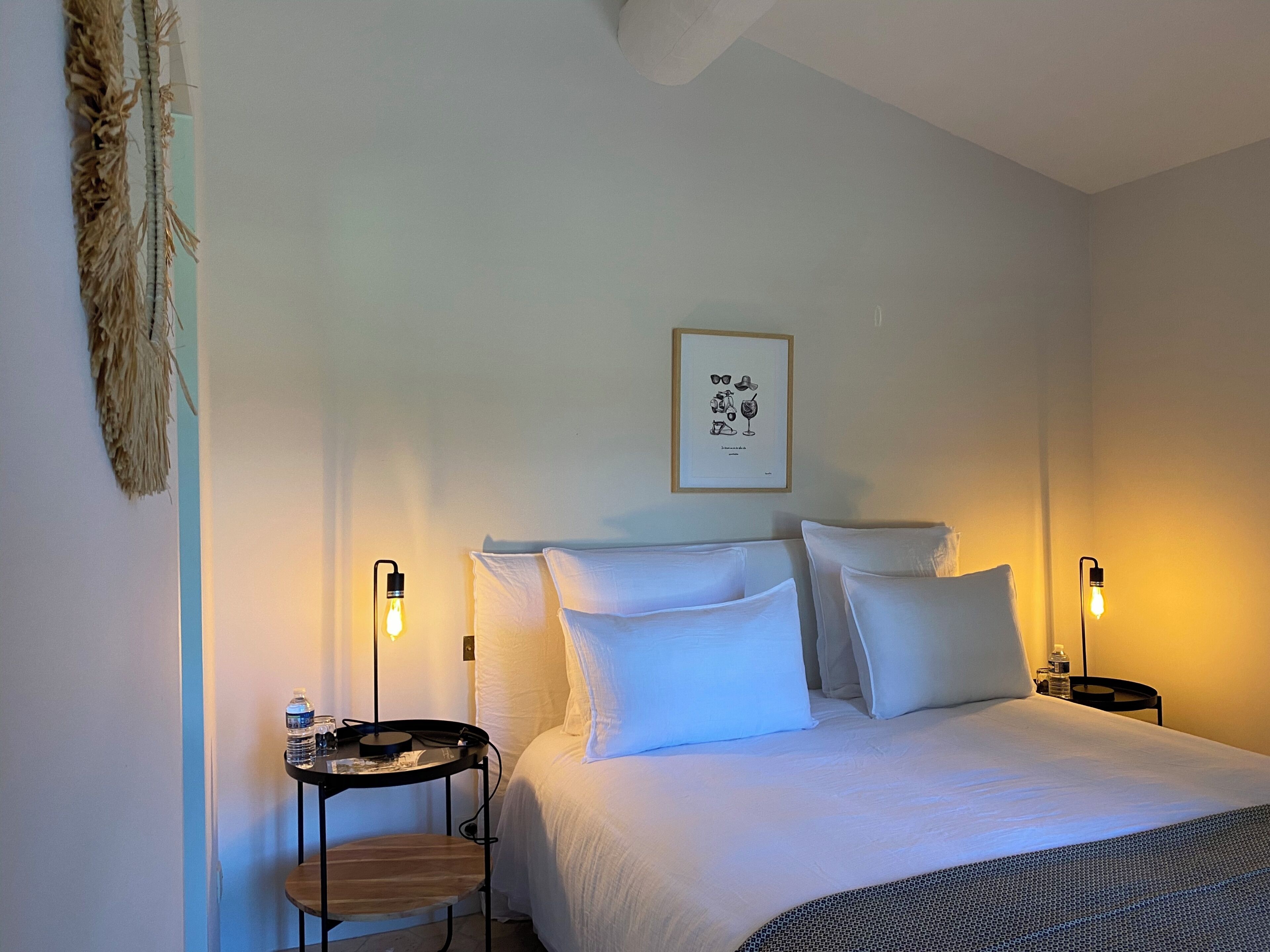 A hotel room with a bed, two bedside tables, a framed picture on the wall, and a hanging decoration.
