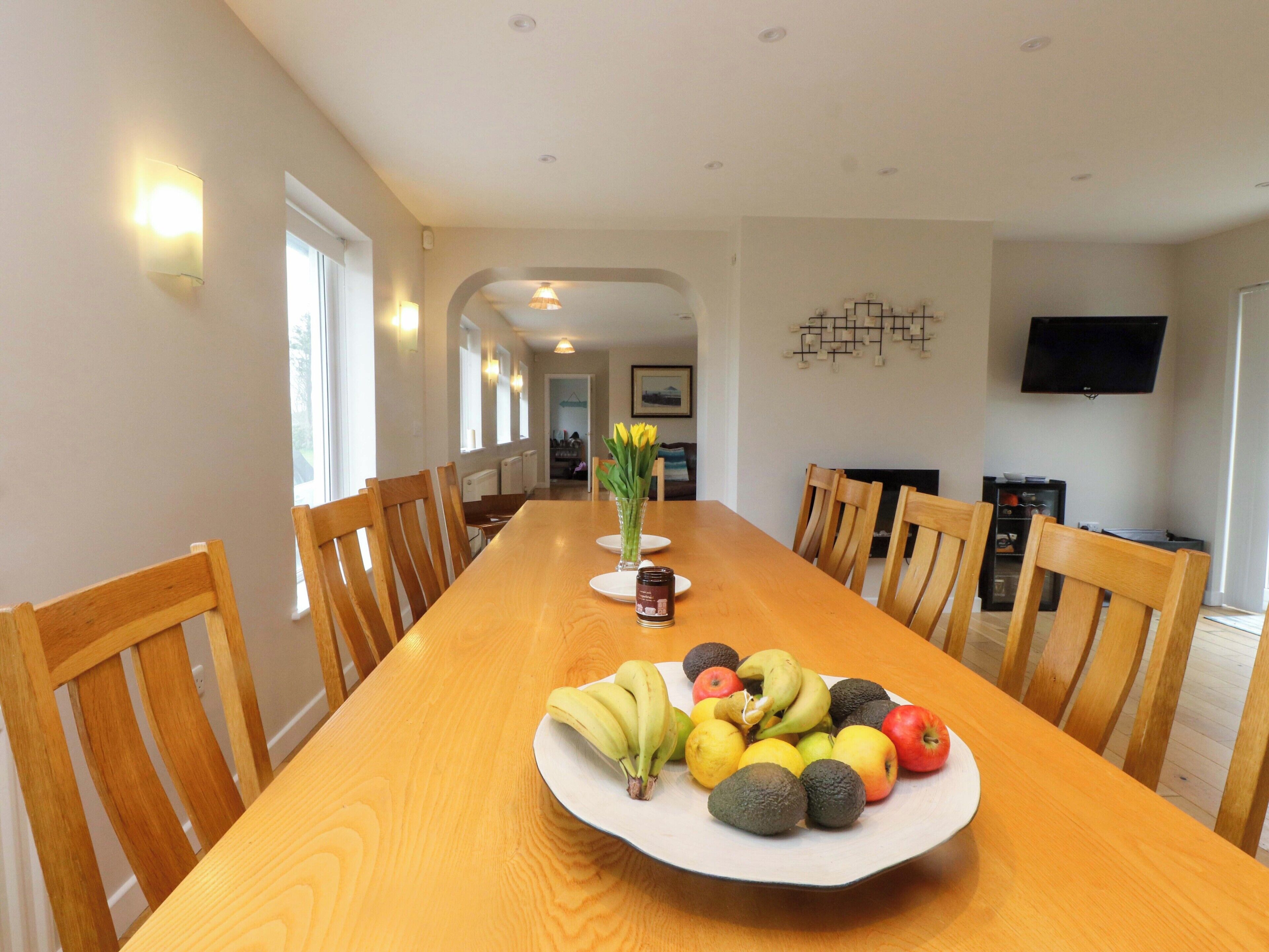 A dining area with a long wooden table, chairs, a fruit plate, and a vase with flowers.