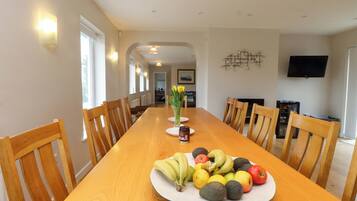 A dining area with a long wooden table, chairs, a fruit plate, and a vase with flowers.