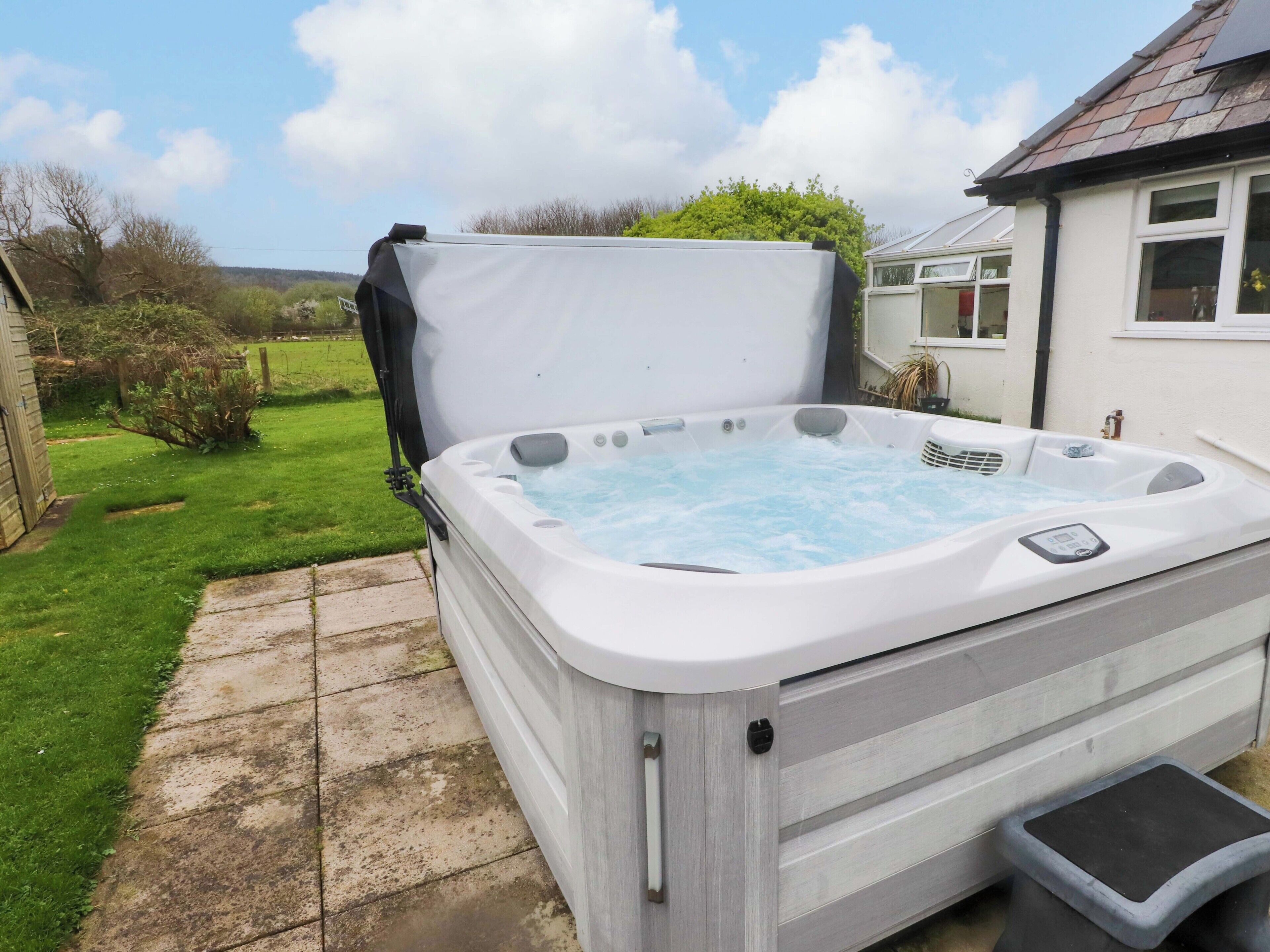 Un jacuzzi con tapa, un taburete y un jardín con césped y una casa al fondo.