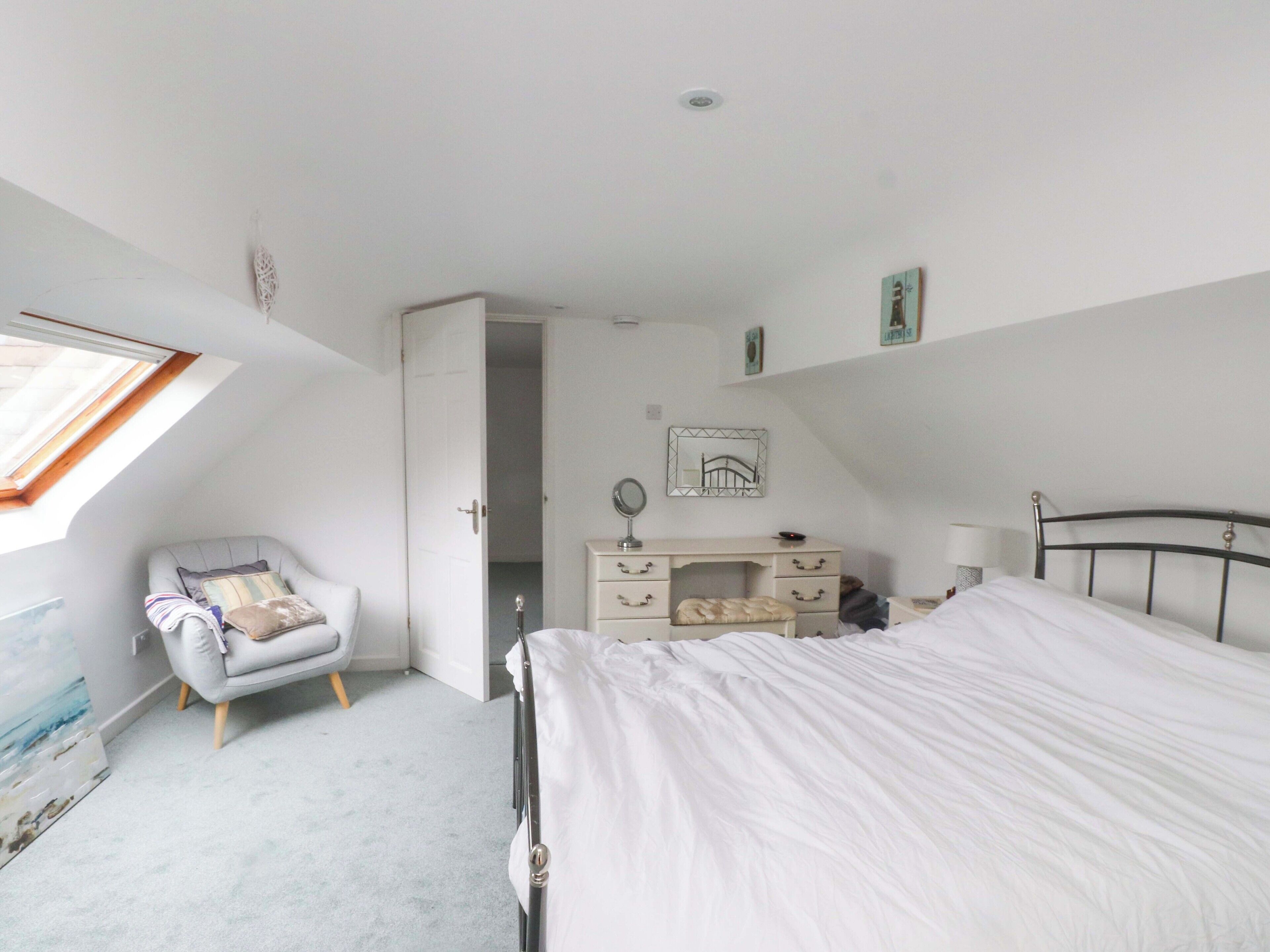 Un dormitorio con cama, cómoda, silla, escritorio, espejo y tragaluz.