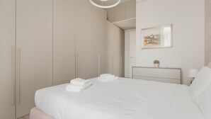 Ein modernes Schlafzimmer mit einem großen Bett, einem Nachttisch mit Lampe, einem kleinen Topfpflanzen und einem gerahmten Bild an der Wand.