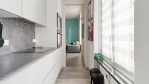 Eine schmale Küche mit weißen Schränken, einer Marmorarbeitsplatte und Blick in einen Wohnbereich mit einer türkisfarbenen Wand und einem Sofa.