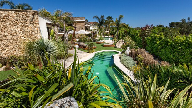 Une piscine entourée d’une végétation luxuriante, de murs en pierre et de palmiers.