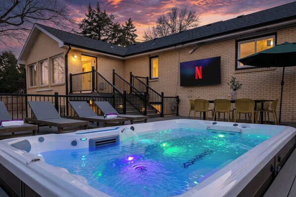A hot tub with a waterfall feature, a TV screen, outdoor seating, and a staircase leading to a covered area.