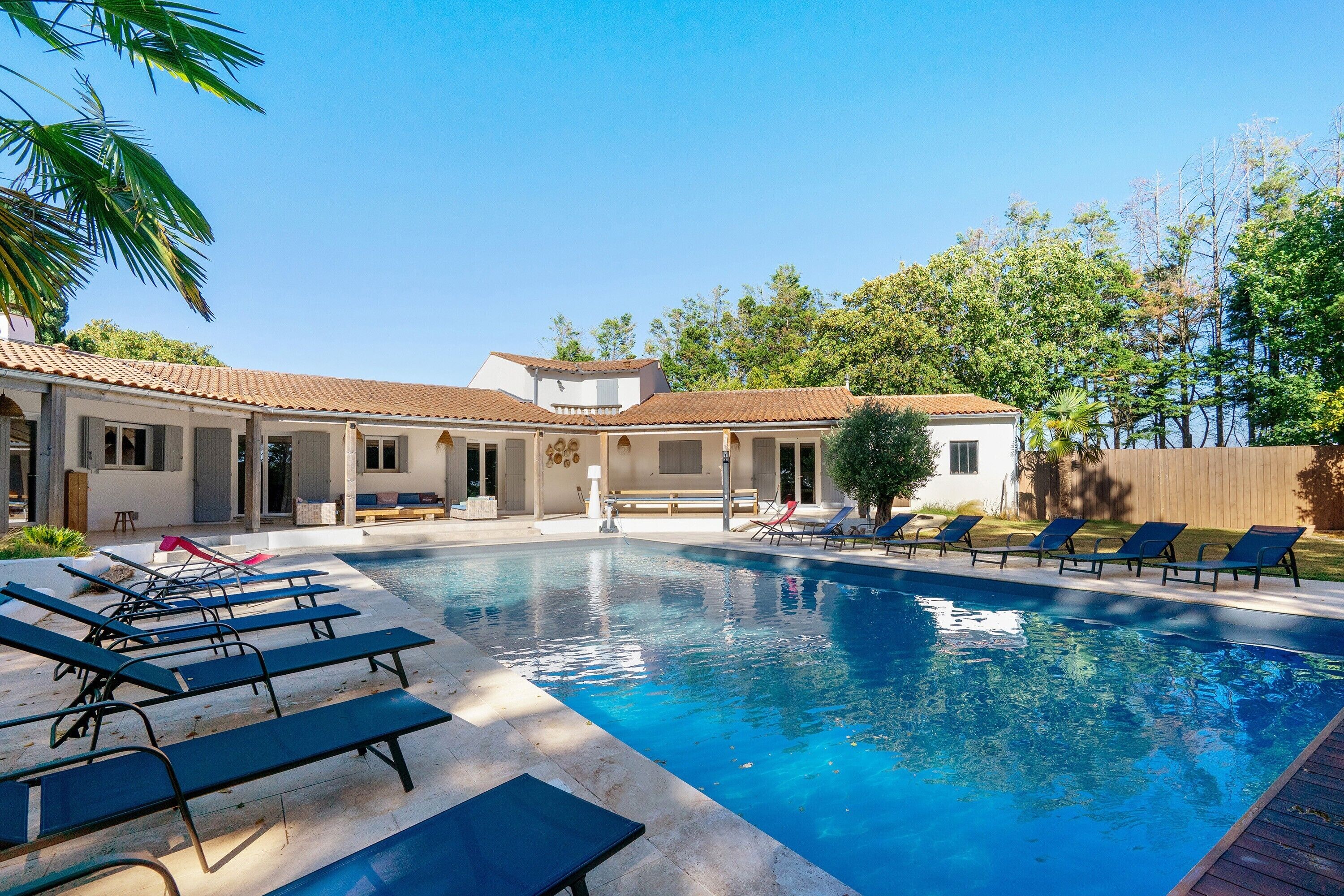 A swimming pool with lounge chairs, a building with a tiled roof, and trees in the background.