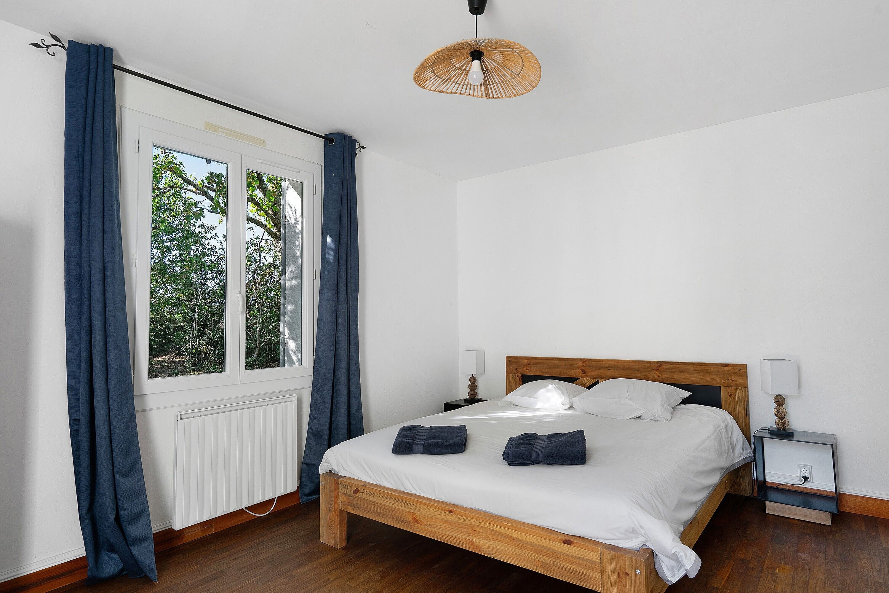 Un dormitorio con cama de madera, ropa de cama blanca, una mesita de noche con lámpara y una ventana con cortinas.