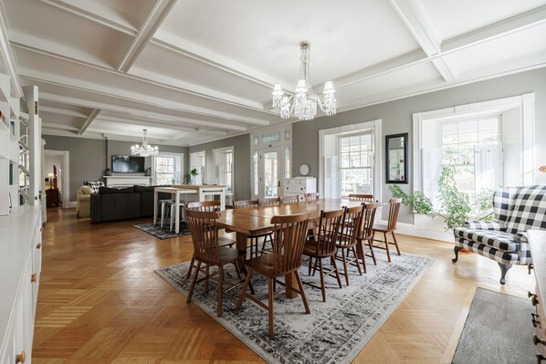 A spacious dining area with a large wooden table and chairs, a chandelier, and a view of the kitchen in the background. - Charming 5-BR estate in FLX perfect for relaxing stays. HOT TUB. GAME ROOM. POOL (Canandaigua)