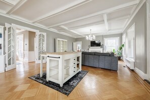 A spacious living room with a wooden floor, a grey sofa, a white entertainment unit, and a chandelier. - Charming 5-BR estate in FLX perfect for relaxing stays. HOT TUB. GAME ROOM. POOL (Canandaigua)