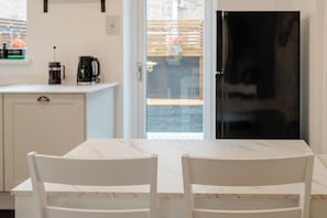 Une table en marbre blanc, accompagnée de deux chaises blanches, une machine à café noire et un meuble blanc, dans une cuisine dont la porte est en verre et donne sur l’extérieur.