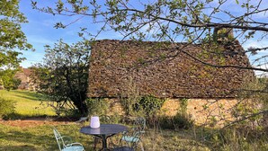 Un bâtiment rustique en pierre, doté d’un toit de chaume, entouré de verdure et agrémenté d’un petit espace extérieur avec des chaises et une table en métal.
