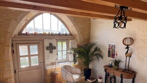 Une pièce chaleureuse avec un plafond en bois, une croix accrochée au mur, une grande fenêtre offrant une belle vue, une chaise, une table avec des livres et une plante.