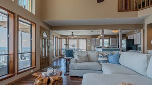 A spacious living room with a large gray sofa, wooden coffee table, and a view of the ocean through large windows. - Stunning Bayfront Home!  Panoramic Views, Fishing, Crabbing and Epic Sunsets. (Jamaica Beach)
