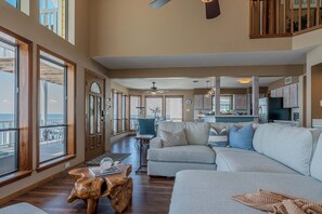 A spacious living room with a large gray sofa, wooden coffee table, and a view of the ocean through large windows. - Stunning Bayfront Home!  Panoramic Views, Fishing, Crabbing and Epic Sunsets. (Jamaica Beach)