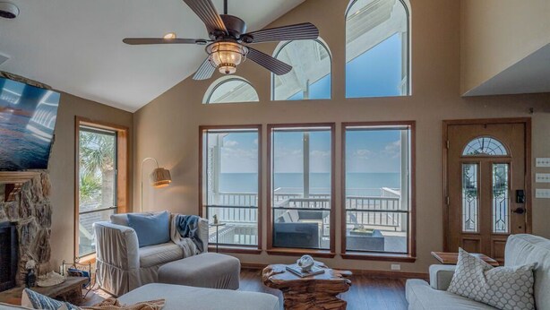 A spacious living room with a high ceiling, large windows, a ceiling fan, and a stone fireplace. - Stunning Bayfront Home!  Panoramic Views, Fishing, Crabbing and Epic Sunsets. (Jamaica Beach)