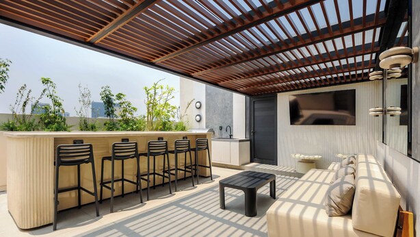 Een moderne buitenruimte met een houten pergola, een bar met krukken, een bank en een aan de muur bevestigde televisie.