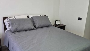 En snyggt bäddad säng med ett grått överkast och kuddar, ett sängbord med en lampa och en enkel, vit vägg.