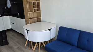 Ett litet, modernt rum med ett runt matbord, en blå soffa, en bokhylla i trä och en liten köksdel.