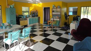 Ein Zimmer mit Schachbrettmuster-Boden, einer Küchenzeile inklusive Kühlschrank, Mikrowelle und Schränken sowie einem Essbereich mit Tisch und Stühlen.