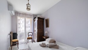 Ein Schlafzimmer mit Bett, Sessel, Kleiderschrank, Lampe und einem Fenster mit Vorhängen.