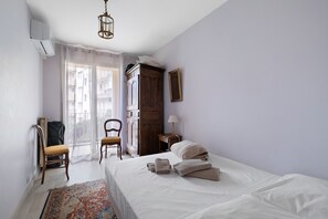 A bedroom with a bed, a chair, a wardrobe, a lamp, and a window with curtains. - Central Juan les Pins – Spacious 3-Bedroom Sea View Apartment (Antibes)