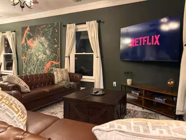 Un salon avec un canapé en cuir, une table basse, un téléviseur à écran plat affichant le logo « NETFLIX », et un grand tableau abstrait accroché au mur.
