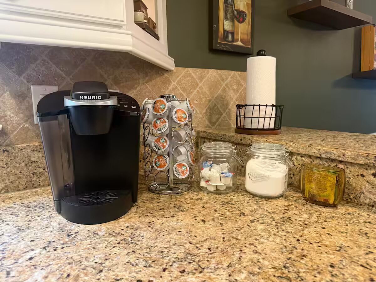 A coffee maker, a jar of coffee pods, a jar of sugar, and a small container on a kitchen counter.