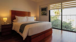 Une chambre à coucher avec une tête de lit en bois, un grand lit recouvert de draps blancs, une table de chevet avec une lampe et une fenêtre donnant sur des palmiers.