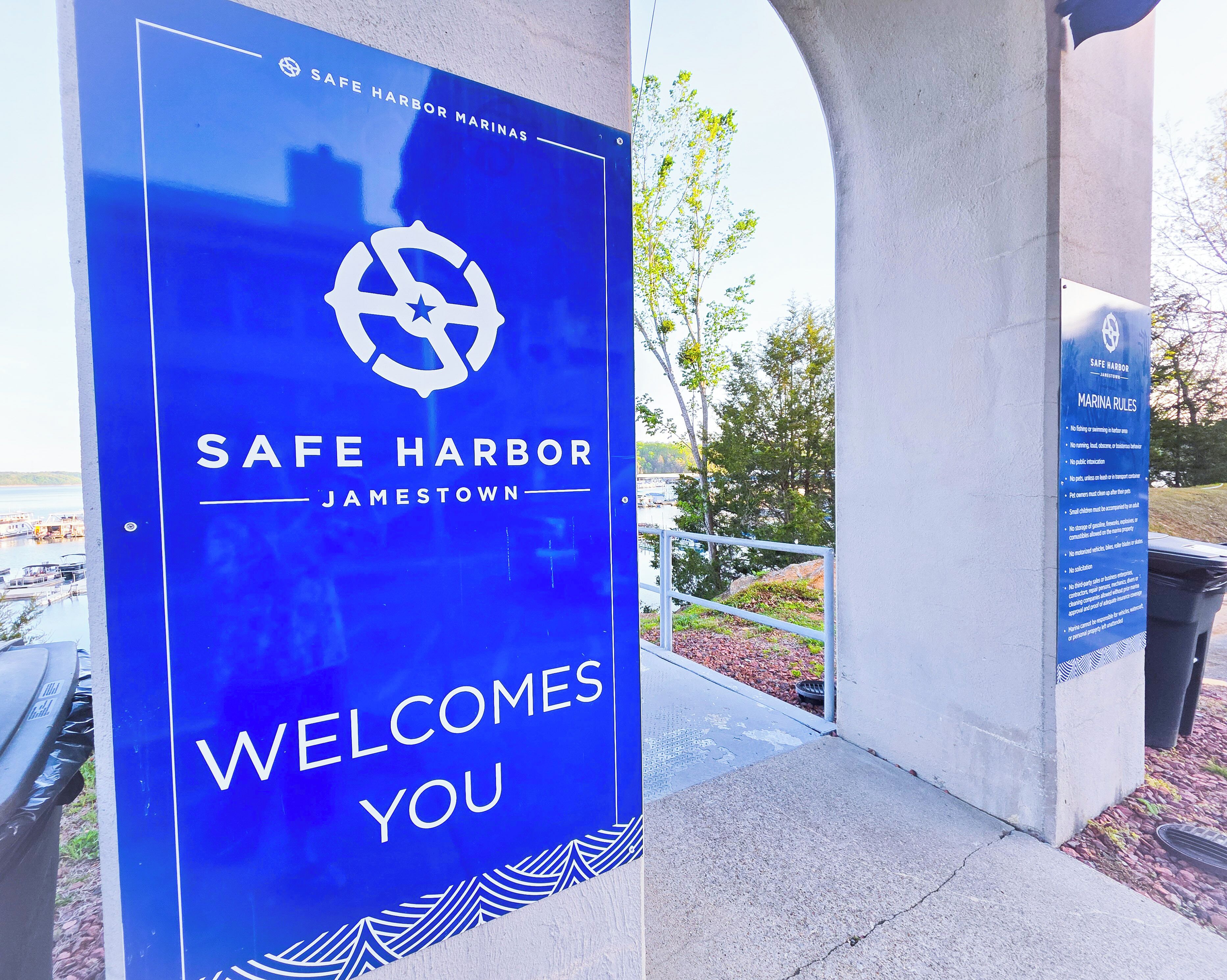 A blue sign with 'SAFE HARBOR JAMESTOWN' and 'WELCOME YOU' text, a marina in the background, and a concrete structure.