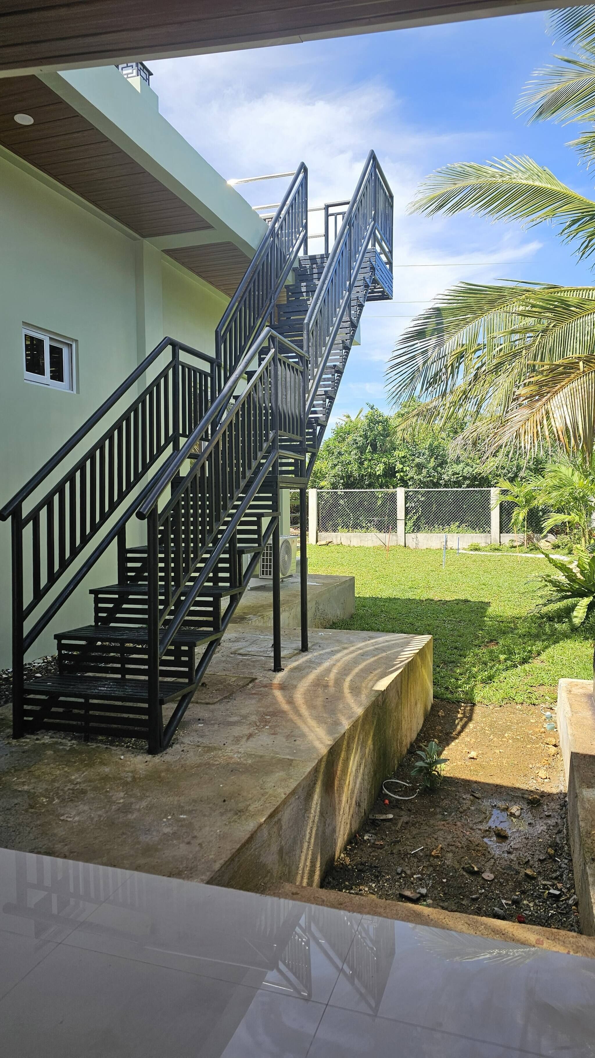 A staircase leading to a building with a green exterior, a concrete base, and a fenced yard with palm trees.