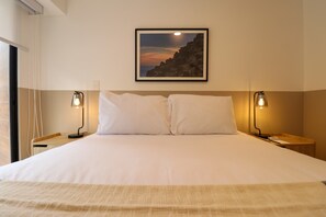 A neatly made bed with two pillows, a framed picture of a sunset on the wall, and two bedside lamps. - w* | Comfortable Living 2BR w/ Balcony in San Isidro (San Isidro)