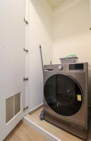 A compact laundry room with a front-loading washing machine, a broom, and a laundry basket on top. - w* | Comfort Living 2BR w/ Balcony in San Isidro (San Isidro)