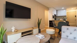 Ein modernes Wohnzimmer mit Fernseher, einem Holz-Konsolentisch, einem runden Couchtisch, einem grauen Sofa und einem Essbereich mit Glastisch und Stühlen.