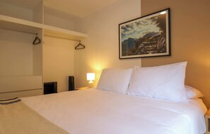 A hotel room with a bed, a nightstand, a lamp, a coat rack, and a framed picture on the wall. - w* | Comfort Living 2BR w/ Balcony in San Isidro (San Isidro)