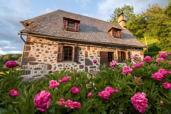 Une maison en pierre avec un toit en pente, des volets en bois et un jardin où poussent des fleurs roses.
