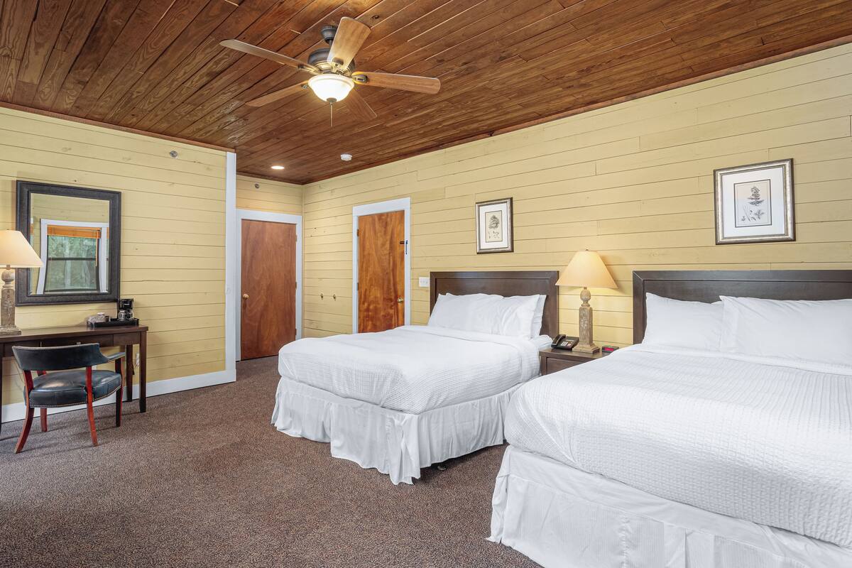 A room with two beds, a desk, a chair, a ceiling fan, and framed pictures on the walls.