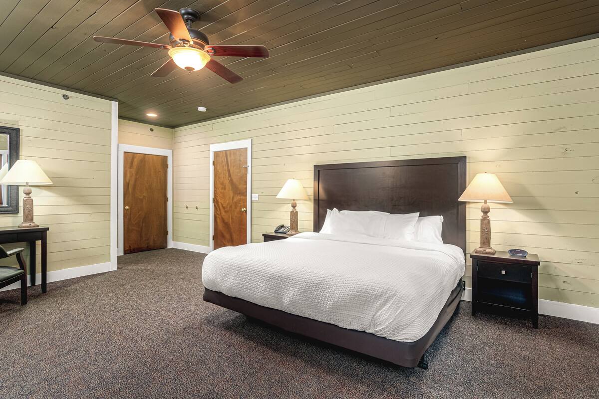 A bedroom with a large bed, two bedside tables with lamps, a ceiling fan, and wooden doors.