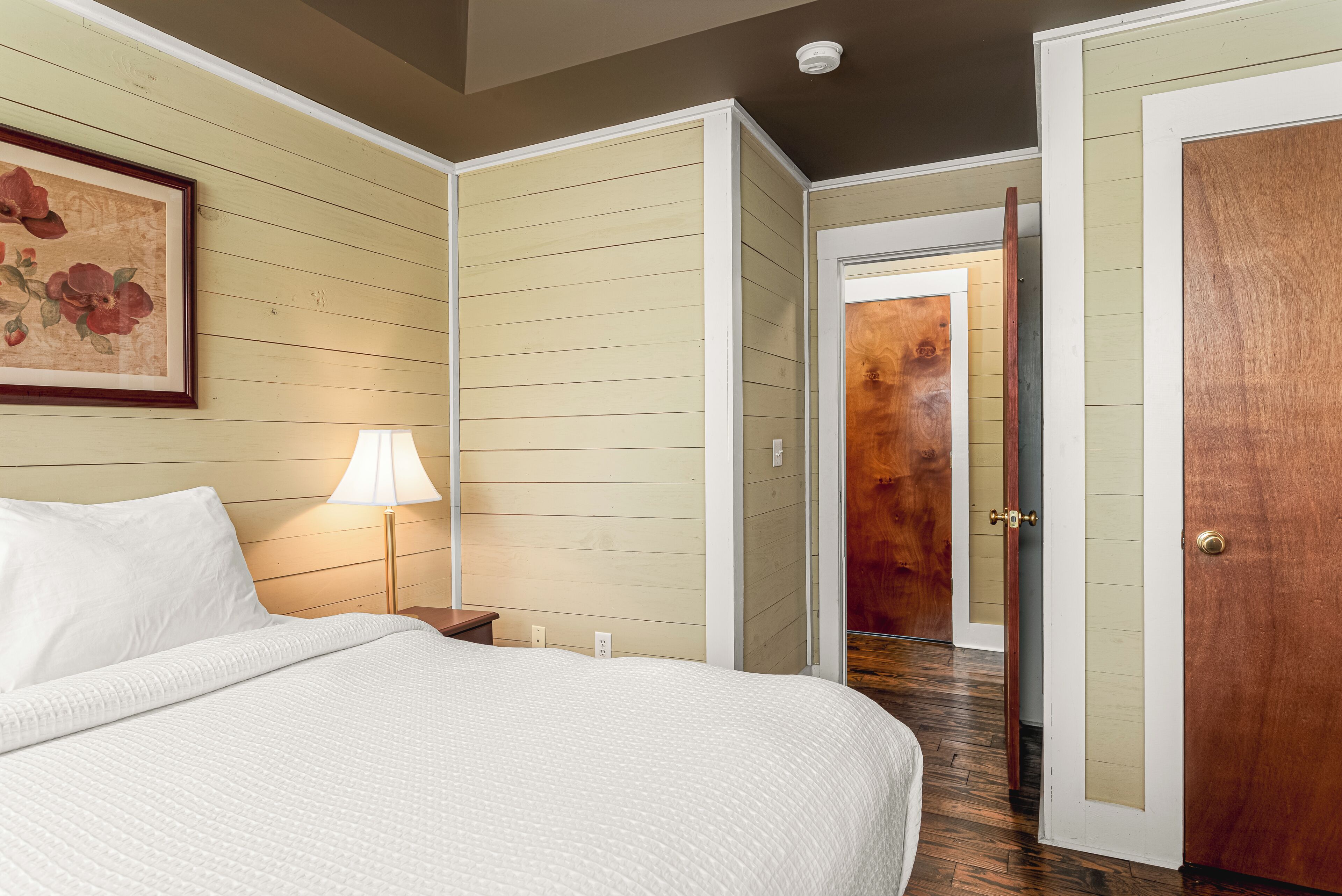 A hotel room with a bed, a nightstand, a lamp, a framed picture, and a wooden door.