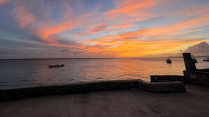 Una puesta de sol sobre un mar tranquilo con barcos, un muro de piedra y una playa arenosa.