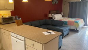 Una kitchenette compacta con tostadora, pileta y encimera. Se observa una cama con frazada y almohadas coloridas, un sofá azul y un aire acondicionado instalado en la pared.