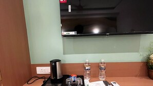 A hotel room with a flat-screen TV mounted on the wall, a wooden desk with a coffee maker, two glasses, two bottles of water, a remote control, and a trash bin.