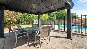 A covered patio with a glass table and chairs, a pool in the background, and a fenced yard. - Tranquil poolside retreat!  Basement with 2 rooms! Close to Niagara Falls. (Niagara Falls)