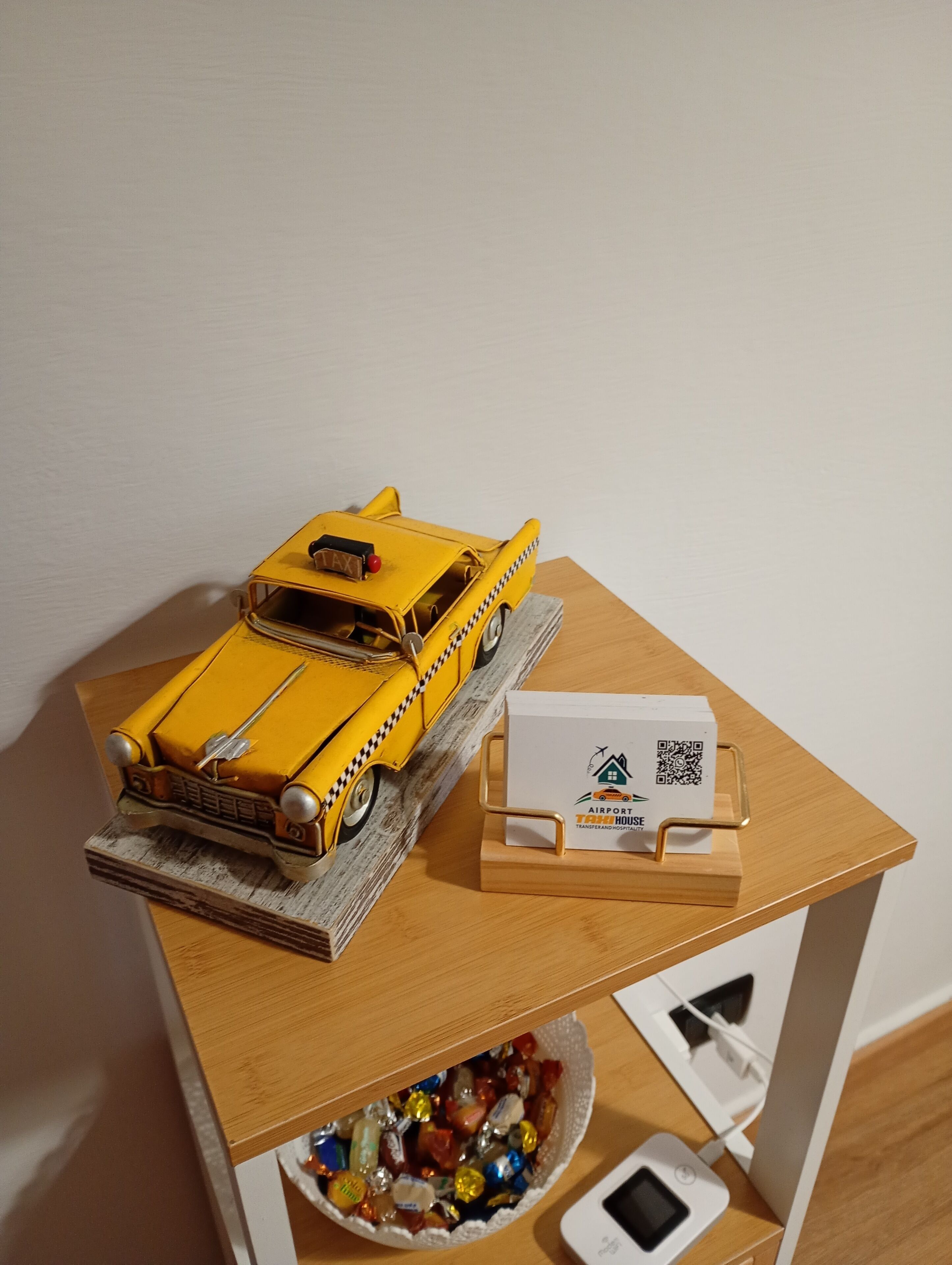 Ein gelbes Modelltaxi auf einer Holztischplatte, eine weiße Schachtel mit einem QR-Code und eine Schale mit bunten Bonbons.