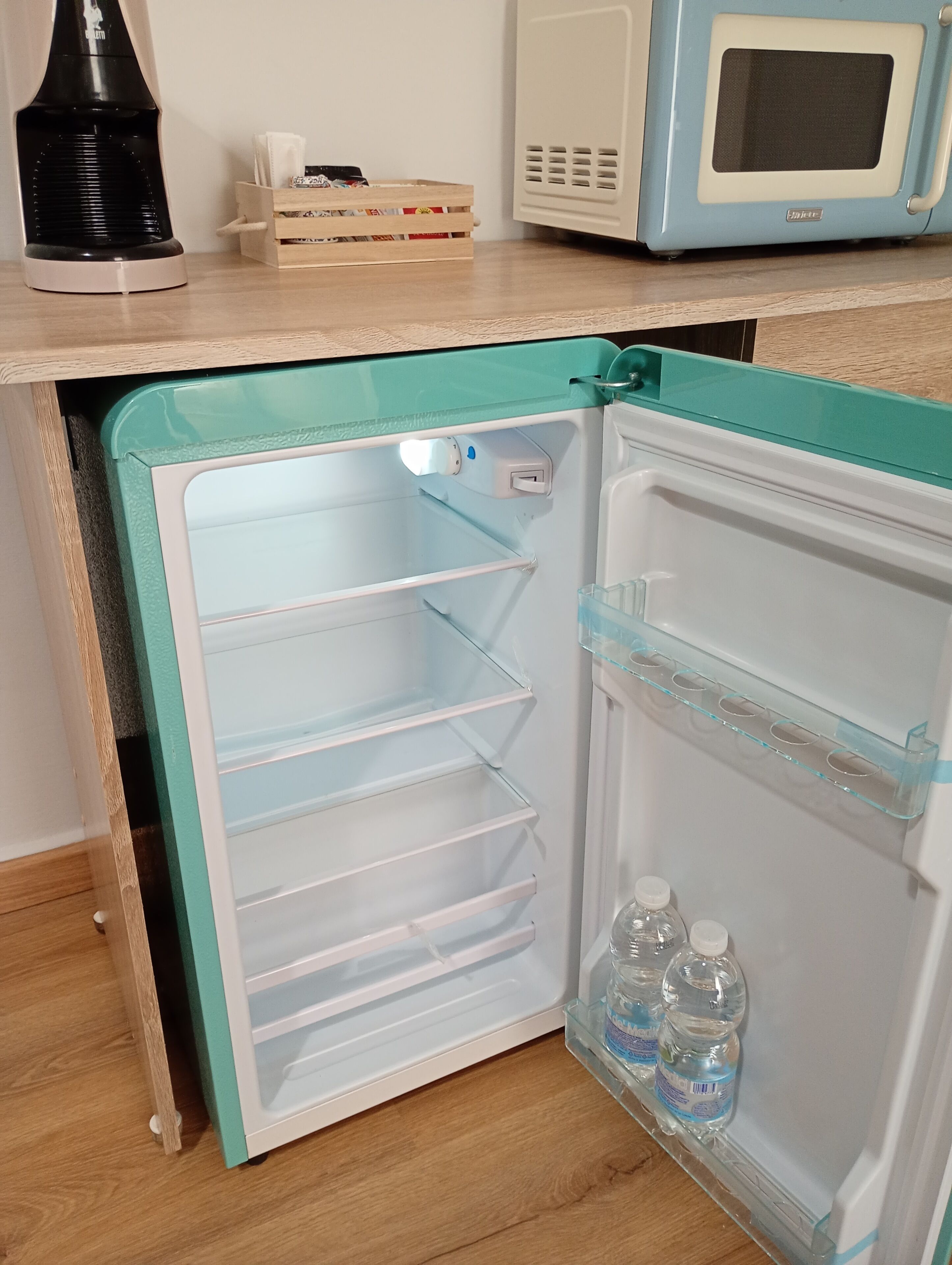 Ein kleiner Kühlschrank mit türkisblauem Äußeren, einem Gefrierfach und einer Ablage mit Wasserflaschen.
